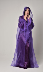 full length portrait of beautiful girl with long black hair,   wearing purple fantasy medieval gown and cloak. standing pose on grey studio background.