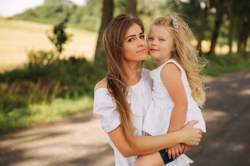 Attractive Mom and blonde hair daughter sits on road near big alley. They smile and look to natune. Front view