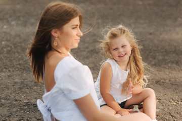Fototapeta na wymiar Attractive Mom and blonde hair daughter sits on road near big alley. They smile and look to natune