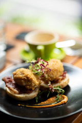 Eggs Benedict with fried breadcrumb eggs in toasted buns, with bacon and salad in a black dish.