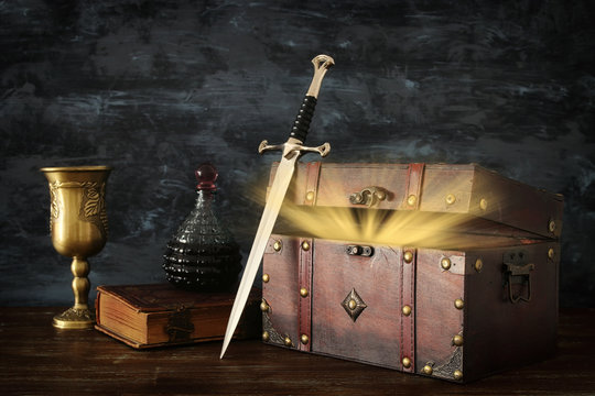 low key image of open chest with treasure, wine cup, antique old book and sword. fantasy medieval period.