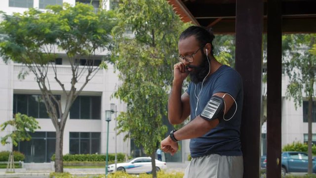 Medium Shot of young Indian man standing and checking gadgets after jogging during sunny day in urban area