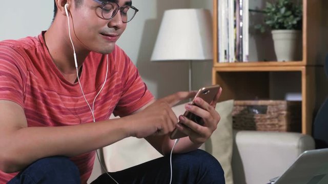 Medium shot of young Asian man in colorful stripped t-shirt sitting in apartment, looking at pictures in phone and listening to music through headphones