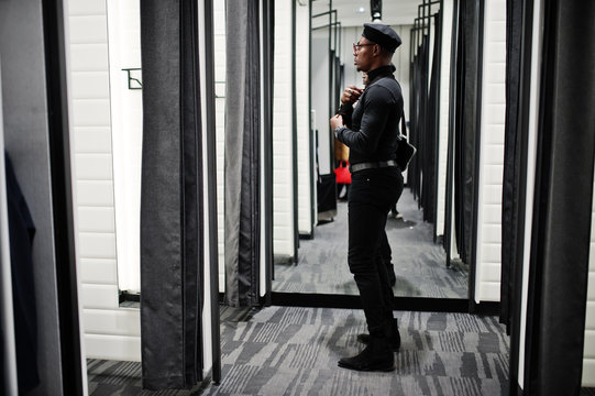 Stylish casual african american man at black outfit and beret with waist bag at fitting room clothes store , looking at mirror.