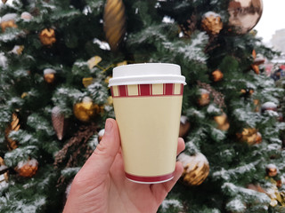 Tea or coffee in disposable cup in hand on Christmas Eve on the background of Christams tree with decorations