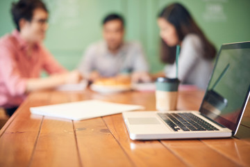 Wooden table with laptop and modern colleagues having coffee and working on background