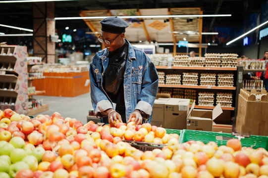 Stylish casual african american man at jeans jacket and black beret checking apple fruits in organic section of supermarket.