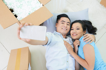 Young romantic Asian couple taking selfie while lying on floor in new apartment with carton boxes around