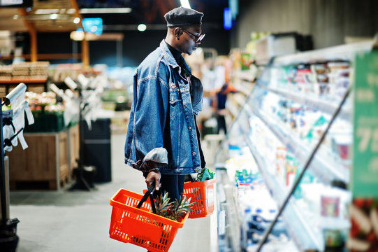 Stylish casual african american man at jeans jacket and black beret holding two baskets, standing near fridge and shopping at supermarket.