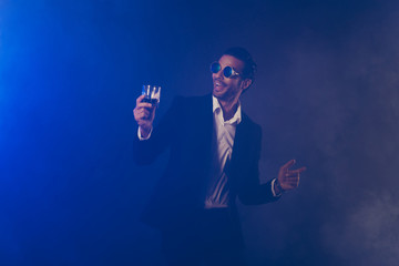 Portrait of nice attractive handsome cheerful cheery glad man wearing black jacket eyewear eyeglasses holding glass having fun isolated over dark blue fume smoke background