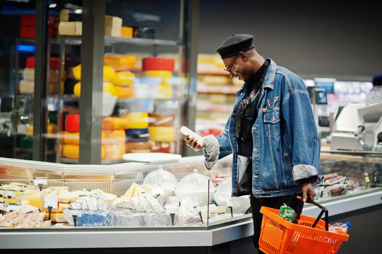 Stylish casual african american man at jeans jacket and black beret holding basket, standing near cheese fridge and shopping at supermarket.