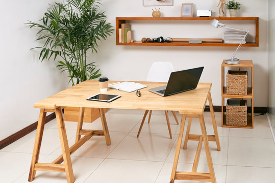Empty wooden office table with laptop, touchpad and notepad on it with green plant and big shelf in the background
