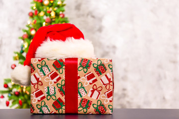Festive composition with one single present in colorful wrapping paper with santa claus hat on foreground and blurry decorated Christmas spruce tree on background. Close up, copy space.