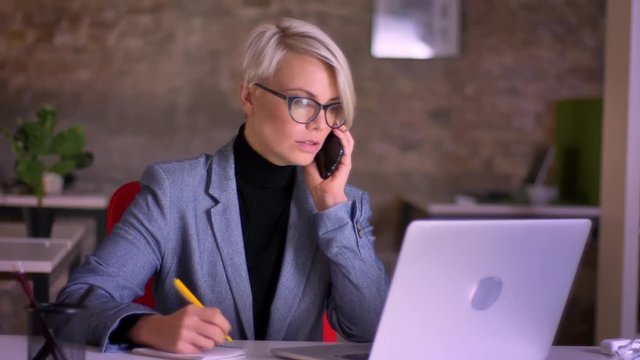 Blonde short-haired businesswoman in glasses talking on phone, working with the laptop and making notes in office.
