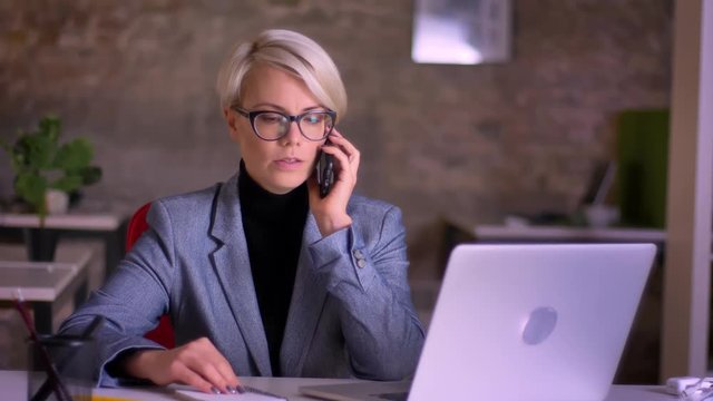 Portrait of blonde short-haired businesswoman in glasses seriously talking on cellphone in office.