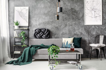 Dark green blanket on comfortable grey sofa in elegant industrial living room with maps on empty...