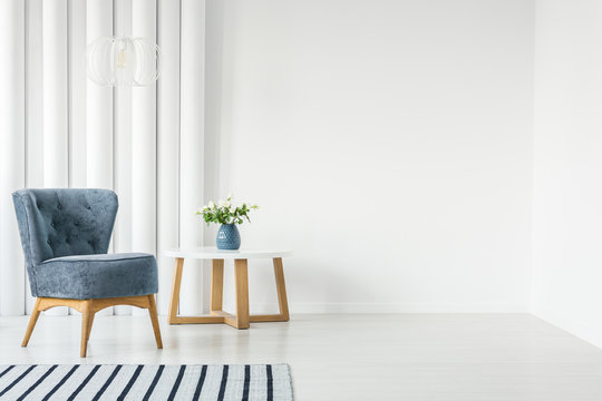 Fashionable blue armchair next to white round coffee table with vase with flowers in empty white interior with striped rug
