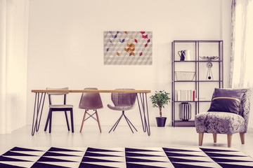Pillow on grey armchair and geometric carpet in bright dining room with clock and poster on the wall and plant on the floor