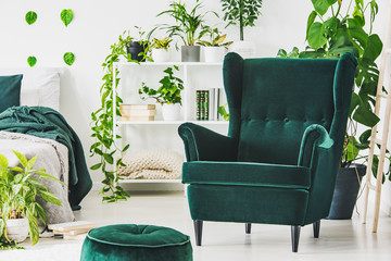Emerald green comfortable armchair and pouf in contemporary bedroom interior with urban jungle