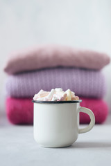 Winter cozy concept. Sweater weather. Coffee with marshmallows in white enameled metal cup on the background of  stack of pink knitted sweaters on table. Warm weekend in cold weather.