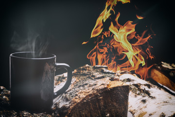 Black cup of hot steaming coffee sitting on an old log by campfire