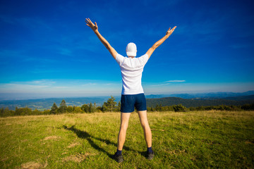 Summer hiking in mountains. Young tourist man in cap with hands up on top of mountains admires nature.