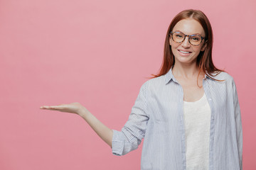 Studio shot of pleased Caucasian woman keeps palm raised, wears optical glasses, demonstrates something over copy space, holds blank space, has pleasant smile on face, poses indoor over pink wall