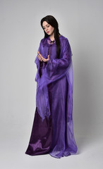 Naklejka premium full length portrait of beautiful girl with long black hair, wearing purple fantasy medieval gown and cloak. standing pose on grey studio background.