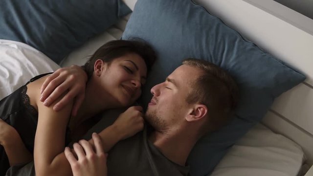 Young caucasian loving couple talk and kiss while lying in bed, waking up in the morning. Top view of attractive man kissing his smiling brunette wife. Slow motion