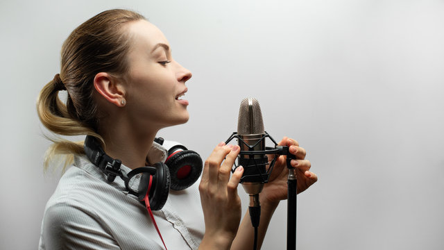 Young beautiful girl writes vocals, radio, voiceover tv, reads poetry, blog, podcast in studio on studio microphone in headphones on white background
