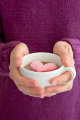 woman with purple pullover holding white cup filled with hot chocolate with a pink heart-shaped marshmallow