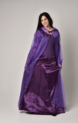 Naklejka premium full length portrait of beautiful girl with long black hair, wearing purple fantasy medieval gown and cloak. standing pose on grey studio background.