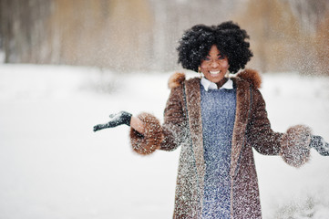 Curly hair african american woman wear on sheepskin coat and gloves posed at winter day throws up snow.