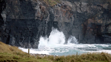 Naklejka premium White waves clashing against a dark rocky cliff, with some grass in the foreground.