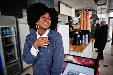 Curly hair african american woman wear on sweater posed at cafe indoor with cup of tea or coffee.