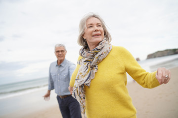 Vibrant senior woman holding husband's hand and leading the way on beach walk