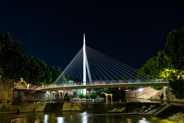 Fototapeta na wymiar Puente colgante uniendo la ciudad que divide en dos el rio. El puente está iluminado y de él cuelgan esculturas y se puede ver el rio y patos