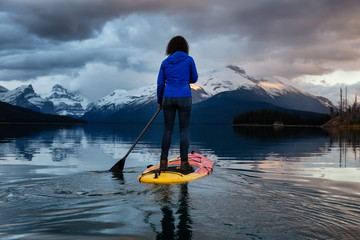 Girl Paddle Boarding in a peaceful and calm glacier lake during a vibrant cloudy sunset. Taken in Maligne Lake, Jasper National Park, Alberta, Canada.