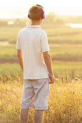 teenager in headphones listening to music on nature, young man is relaxing and looking at sunset in the summer field