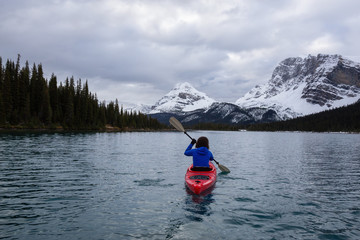 Adventurous girl kayaking in a glacier lake surrounded by the Canadian Rockies during a cloudy morning. Taken at Bow Lake, Banff, Alberta, Canada.