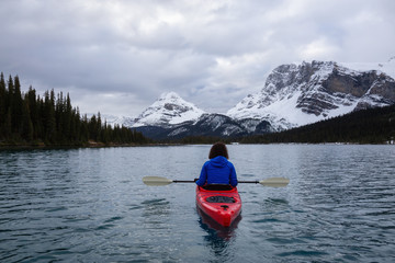 Adventurous girl kayaking in a glacier lake surrounded by the Canadian Rockies during a cloudy morning. Taken at Bow Lake, Banff, Alberta, Canada.