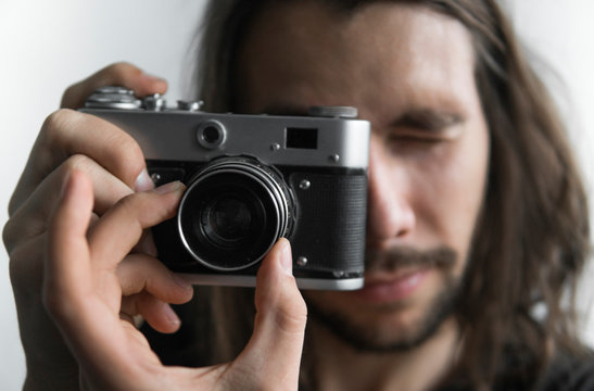 Handsome young bearded man with a long hair and in a black shirt holding vintage old-fashioned film camera on a white background and looking in camera viewfinder.