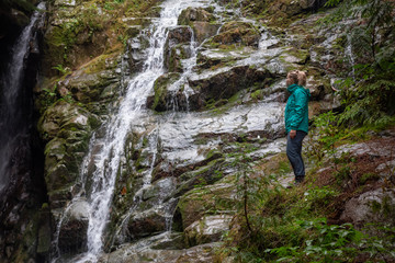 Naklejka na ściany i meble Adventurous female hiker is enjoying a beautiful view of a waterfall during a foggy day. Taken in Mt Fromme, North Vancouver, British Columbia, Canada.