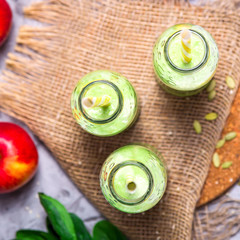 Green smoothies in three glass bottles with tubes on a gray table with a round stand and burlap among apples, spinach and seeds. Top view, flat lay