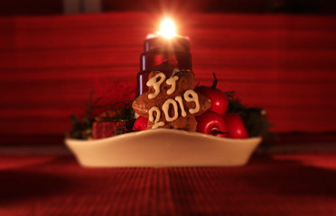 Homemade gingerbread star covered with white icing in form PF 2019 leaning againts fruits. It is in front of candlestick. And first event is behind us