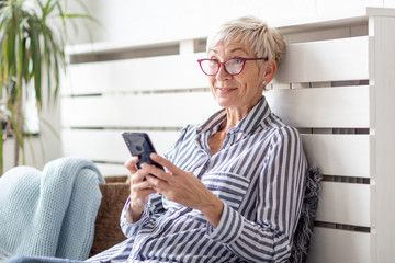 Mature woman with mobile phone on her hands sitting in room and sending messages to her friends and...