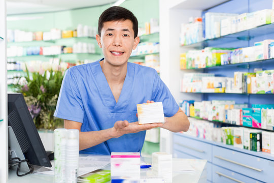 Portrait of chinese man pharmacist who is standing on his work place near cashbox in apothecary.
