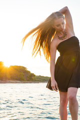 Naklejka premium A beautiful woman in an elegant black dress standing and posing on the rocks next to the sea during the sunset.