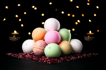 Pyramid of colorful bath bombs on the dark background surrounded by dried rose petals, candles and light in the background