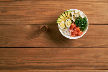 poke bowl with salmon, avocado, rice, Chuka Salad, cheese,, quail eggs sprinkled with white and black sesame isolated on wooden background top view flat lay